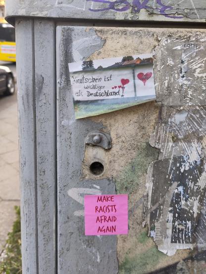 A weathered metal utility box with various stickers and graffiti. On the upper left, there is a sticker with a picture of a house and trees, accompanied by the text "Knutschen ist wichtiger als Deutschland!" (Kissing is more important than Germany!) in German, with red hearts drawn around the text. 
Below this, a pink sticker reads "MAKE RACISTS AFRAID AGAIN" in bold, capitalized letters. 
The box has been exposed to the elements for a considerable time.
In the background is a street scene with a yellow bus (BVG) and a car.