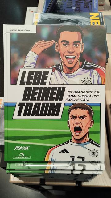 Ein Stapel mit dem Begleitband zur Ausstellung, Titel „Lebe deinen Traum“. Darauf KI-generierte Bilder von Jamal Musiala und Florian Wirtz im Graphic-Novel-Stil.