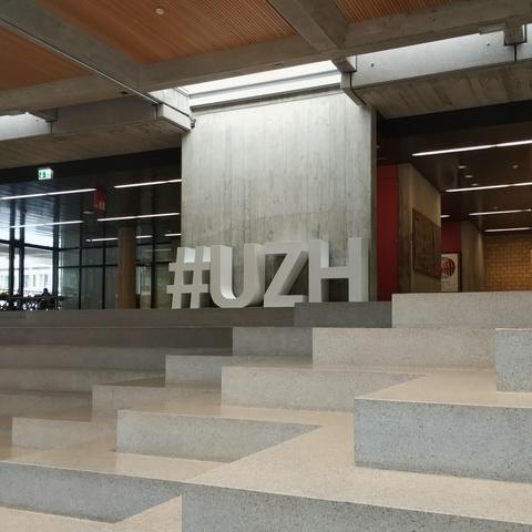 A photo of a hall at university of Zurich showing concrete stairs and large metal letters spelling #UZH.